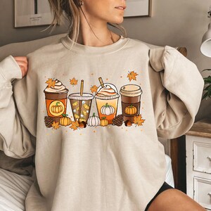 May include: Beige sweatshirt featuring a fall-themed graphic. The design includes four different coffee drinks with pumpkin and leaf accents. Pumpkins, pinecones, and fall leaves are scattered around the drinks. The sweatshirt is a neutral color.