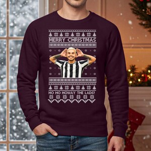 May include: A burgundy Christmas sweater with a festive design. The sweater features the text "MERRY CHRISTMAS" and "HO HO HOWAY THE LADS!" in white. A graphic of a person in a black and white striped shirt is in the center. The sweater has a crew neck.