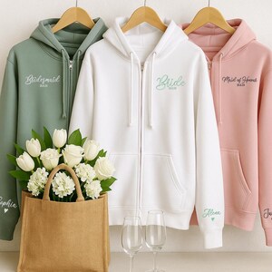 May include: Three zip-up hoodies in sage green, white, and pink, each with custom text. The sage green hoodie says "Bridesmaid", the white hoodie says "Bride", and the pink hoodie says "Maid of Honor". A bouquet of white flowers sits in a burlap tote bag.