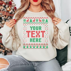 May include: A cream-coloured Christmas jumper with a festive design. The jumper features a red and green pattern with reindeer, Christmas trees, and the text "YOUR TEXT HERE" in a bold, pixelated font. The jumper is a cosy, long-sleeved garment.