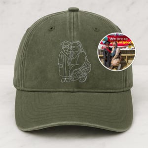 May include: An olive green baseball cap featuring a white outline of a person and a child in graduation robes. A small circular image is on the side. The cap has a curved peak and an adjustable strap at the back.