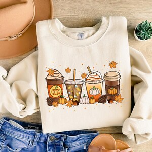 May include: Cream-colored sweatshirt featuring a fall-themed graphic of four coffee drinks with pumpkin designs, surrounded by leaves, pumpkins, and pinecones. The sweatshirt is paired with blue jeans, a brown hat, and sandals.