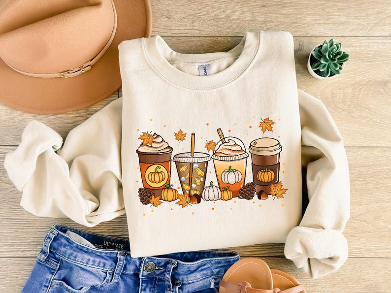 May include: Cream-colored sweatshirt featuring a fall-themed graphic of four coffee drinks with pumpkin designs, surrounded by leaves, pumpkins, and pinecones. The sweatshirt is paired with blue jeans, a brown hat, and sandals.