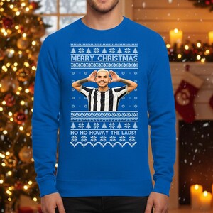 May include: A blue Christmas sweater with a festive design. The sweater features the text "MERRY CHRISTMAS" and "HO HO HOWAY THE LADS!" along with a picture of a person. The sweater has a pattern of snowflakes and Christmas trees.
