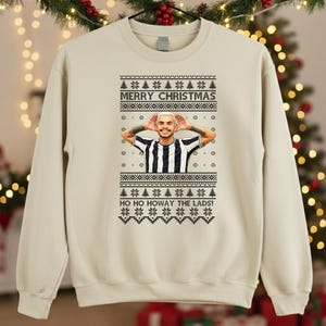 May include: Beige sweatshirt with a festive Christmas design. The design includes the text "MERRY CHRISTMAS" and "HO HO HOWAY THE LADS!" along with a graphic of a person wearing a black and white striped shirt.