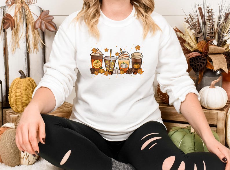 May include: White long-sleeved sweatshirt featuring a colorful graphic of four fall-themed beverages: a pumpkin spice latte, a caramel apple drink, and two other coffee drinks. The design includes pumpkins, leaves, and fall accents.