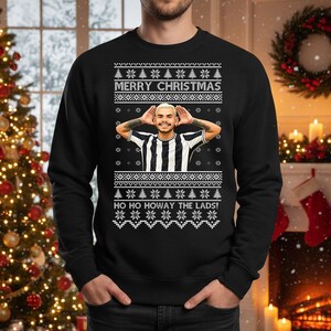 May include: Black Christmas sweater with a festive design. The sweater features the text "MERRY CHRISTMAS" and "HO HO HOWAY THE LADS!" along with a picture of a person. The sweater has a classic crew neck.