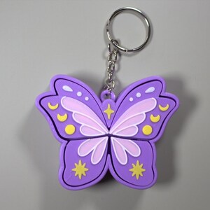 Könnte beinhalten: Ein lila Schmetterling-Schlüsselanhänger mit einem silbernen Schlüsselring. Das Schmetterlingsdesign hat hellviolette und weiße Flügel mit gelben Akzenten. Der Schlüsselanhänger ist aus weichem Material.