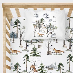 May include: White bedding set with a woodland theme. The set includes a pillowcase and a fitted sheet, both featuring watercolor illustrations of foxes, deer, trees, and forest elements. The design is in shades of brown, green, and gray.