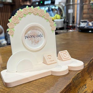 Può includere: Un'insegna da caffè color crema con un design floreale a rose e la scritta "peony café". L'insegna ha una cornice circolare e una base con icone di social media. L'insegna è realizzata in un materiale di colore chiaro.