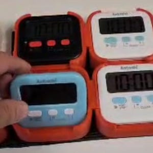 以下が含まれることがあります： 赤、黒、白、青のデジタルキッチンタイマーのコレクション。各タイマーはデジタル画面に「10:00」と表示され、3つのコントロールボタンがあります。タイマーは赤いホルダーに入っています。