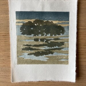 May include: A square woodblock print on textured off-white paper featuring a dark green tree silhouette against a muted blue and beige sky, with a reflective pool of water in the foreground mirroring the tree in shades of green and blue.