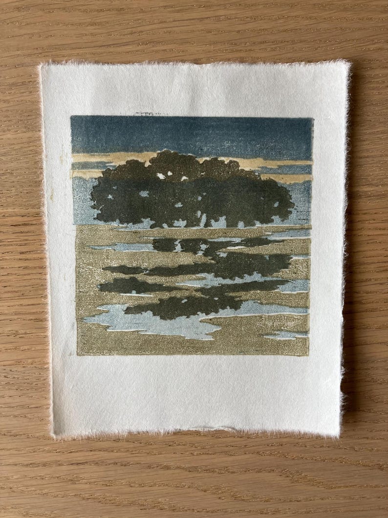 May include: A square woodblock print on textured off-white paper featuring a dark green tree silhouette against a muted blue and beige sky, with a reflective pool of water in the foreground mirroring the tree in shades of green and blue.