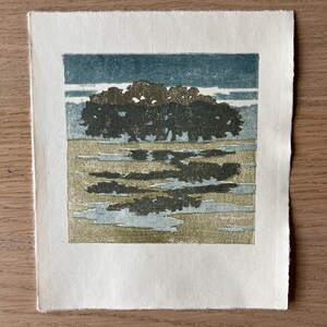 May include: A square block print on off-white paper featuring a dark green cluster of trees against a teal sky. The foreground shows a textured tan field with dark green and light blue reflections, creating a landscape scene with visible ink.