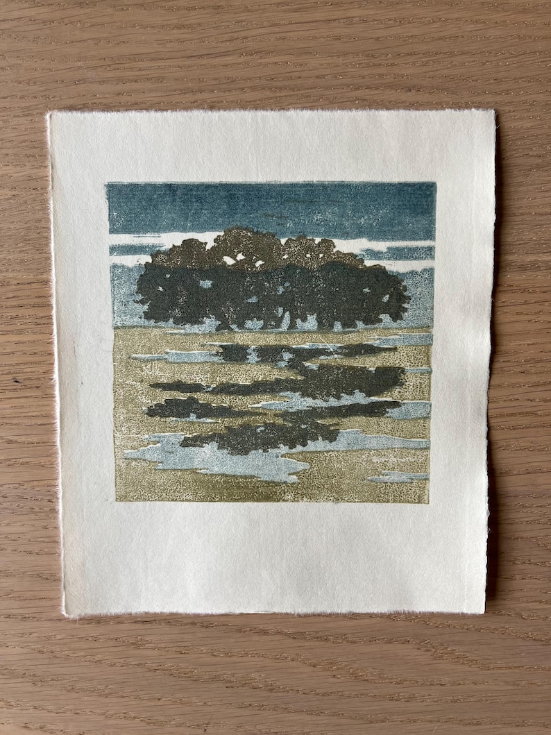 May include: A square block print on off-white paper featuring a dark green cluster of trees against a teal sky. The foreground shows a textured tan field with dark green and light blue reflections, creating a landscape scene with visible ink.