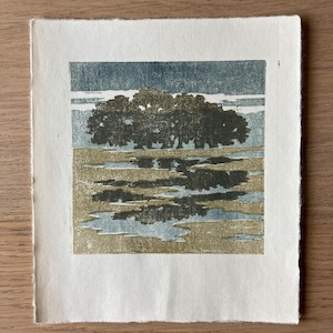 May include: A square block print on off-white paper featuring a dark green cluster of trees centered above a landscape of muted gold and blue horizontal bands. The artwork displays a textured, layered ink application with visible paper edges.