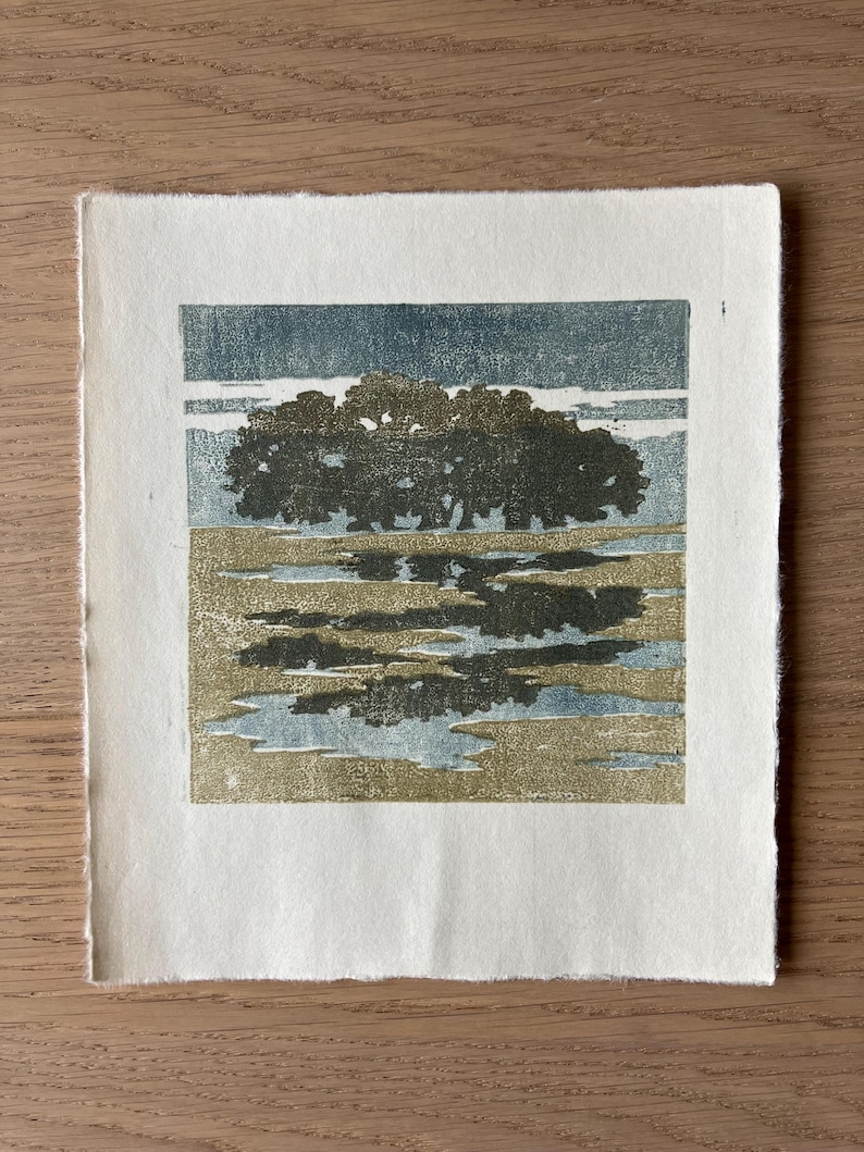 May include: A square block print on off-white paper featuring a dark green cluster of trees centered above a landscape of muted gold and blue horizontal bands. The artwork displays a textured, layered ink application with visible paper edges.