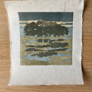 May include: A square block print on textured white paper featuring a landscape with a dark green tree line against a muted blue sky. The foreground shows a reflective body of water in shades of olive green and light blue, creating a calm scene.