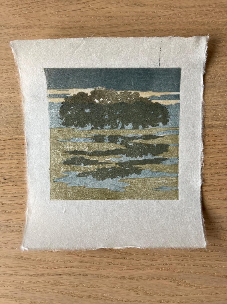 May include: A square block print on textured white paper featuring a landscape with a dark green tree line against a muted blue sky. The foreground shows a reflective body of water in shades of olive green and light blue, creating a calm scene.