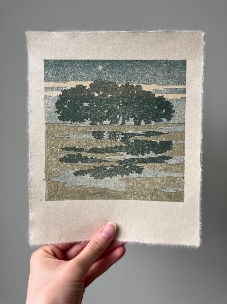 May include: A rectangular block print on textured off-white paper featuring a landscape with a dense cluster of dark green trees under a muted teal sky. The foreground shows abstract patches of green and grey reflecting the tree line.