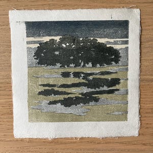 May include: A square woodblock print on textured white paper featuring a dark silhouette of a dense tree line against a muted sky. The foreground displays a landscape with patches of dark shadows and light olive green tones.