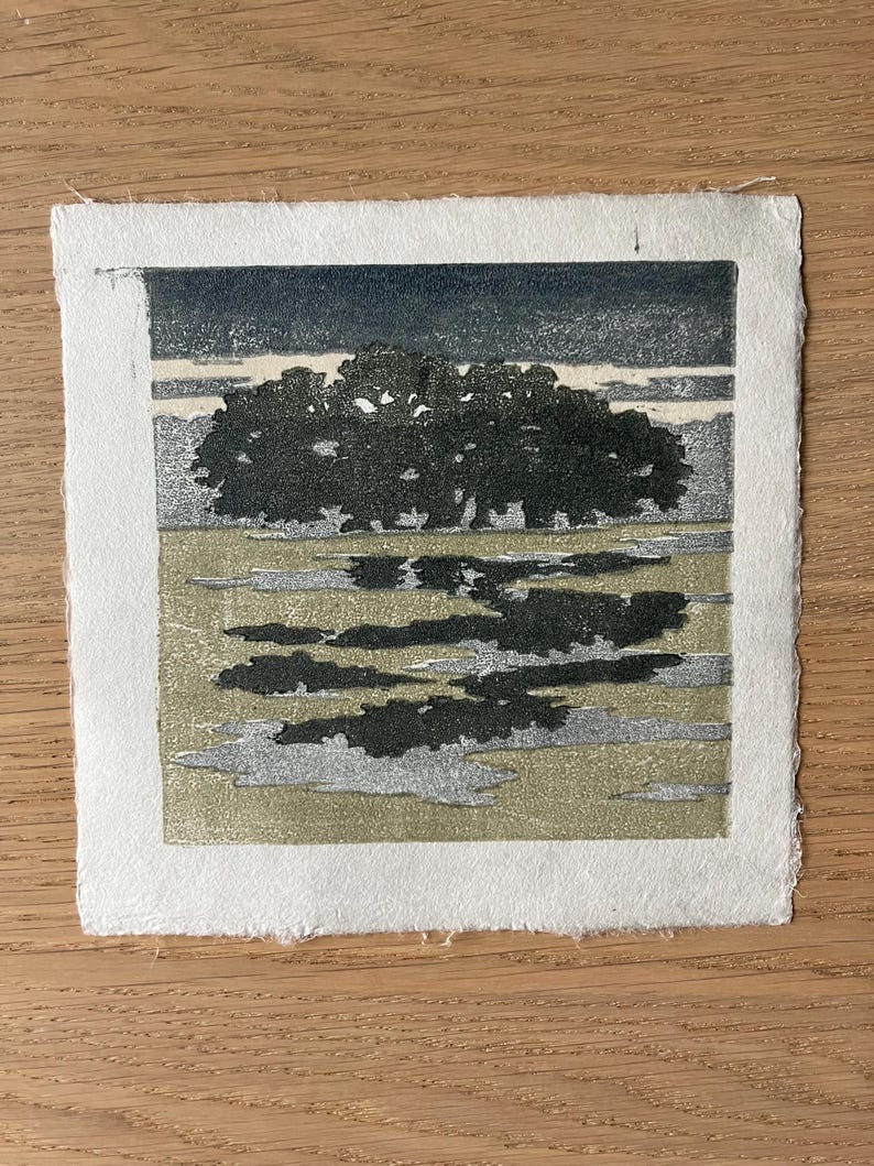 May include: A square woodblock print on textured white paper featuring a dark silhouette of a dense tree line against a muted sky. The foreground displays a landscape with patches of dark shadows and light olive green tones.