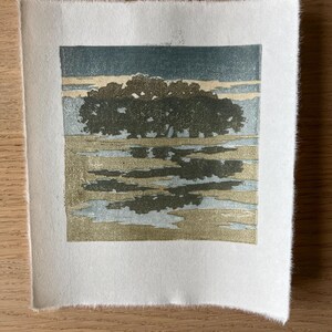 May include: A square block print on textured white paper featuring a landscape with a dark green cluster of trees, a golden yellow field, and a light blue sky. The image shows a reflection of the trees in the foreground water.