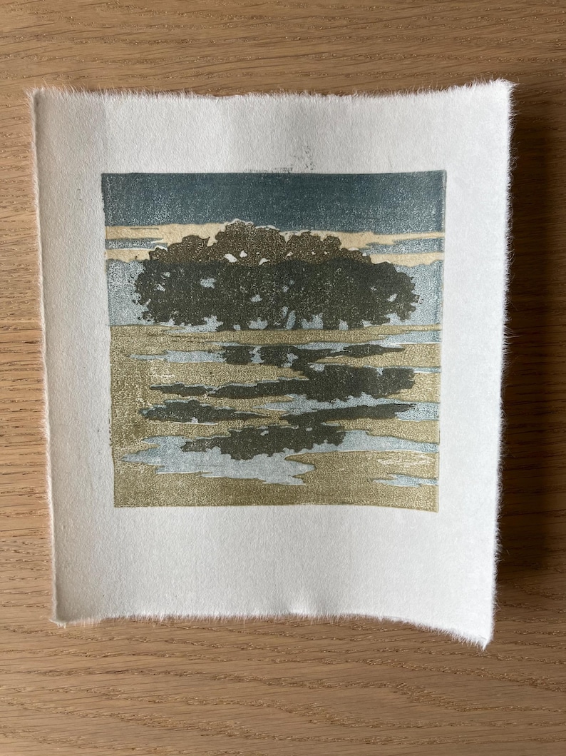 May include: A square block print on textured white paper featuring a landscape with a dark green cluster of trees, a golden yellow field, and a light blue sky. The image shows a reflection of the trees in the foreground water.