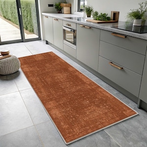 May include: A rectangular runner rug featuring a distressed, burnt orange surface with a subtle, faded pattern. The rug has a thin, light-colored border and is positioned lengthwise on a gray tiled floor in a kitchen setting.