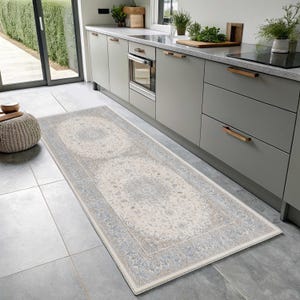 May include: A rectangular runner rug featuring a cream-coloured centre with intricate light blue and grey floral medallion patterns, bordered by a matching detailed frame, laid flat on a grey tiled kitchen floor.