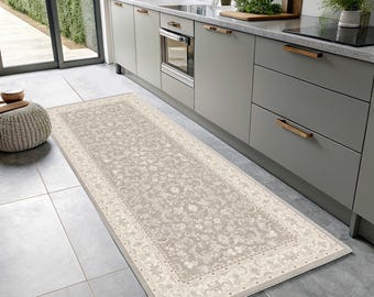 May include: A rectangular, beige kitchen runner rug with an ornate floral pattern and a cream-colored border. The rug is placed on a tiled floor in front of a kitchen counter and cabinets.