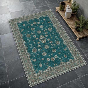 May include: A rectangular area rug featuring a deep teal centre with an intricate floral and vine pattern in shades of cream, beige, and light brown. The rug has a wide, decorative border in muted earth tones with a repeating botanical motif.