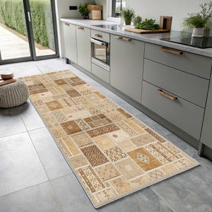 May include: A long rectangular runner rug featuring a patchwork design of various geometric and floral patterns in shades of beige, tan, and brown. The rug is positioned on a gray tiled floor in a kitchen with gray cabinets and a light countertop.