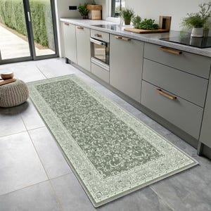 May include: A rectangular, sage green kitchen runner rug featuring an ornate floral design and a cream border. The rug is positioned in front of kitchen cabinets and a counter.