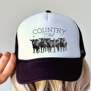 May include: Black and white trucker hat with a white mesh back. The front of the hat features a black and white illustration of cows with a fence and the text "COUNTRY Club".