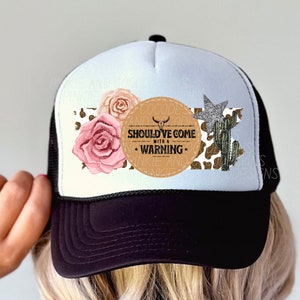 Puede incluir: Gorra de camionero en blanco y negro con un panel frontal blanco con un diseño floral y de temática occidental. El diseño incluye rosas rosas, un cactus, una estrella y el texto "COCODE AND CO. SHOULD'VE COME WITH A WARNING".