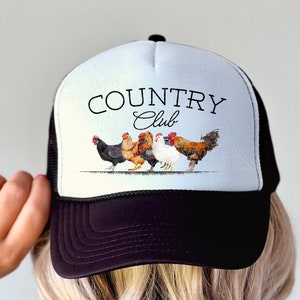 May include: Black and white trucker hat with a white mesh back. The front of the hat features a white design with the text "COUNTRY Club" and a line of colorful chickens.