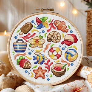 May include: A round cross-stitch embroidery hoop featuring a colorful beach-themed design. The design includes a popsicle, fish, seashell, starfish, and other summer elements. The embroidery is set in a wooden hoop.
