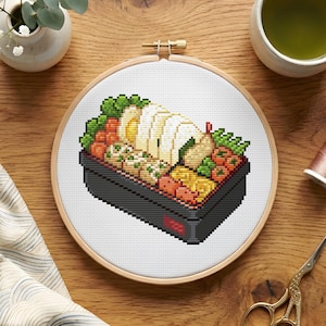 May include: A cross-stitch embroidery hoop featuring a pixelated image of a Japanese bento box. The bento box is filled with various food items in shades of white, orange, yellow, and green. The embroidery hoop is made of wood and is set on a wooden surface.