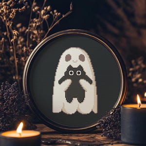 May include: A framed cross-stitch depicting a white ghost hugging a black cat against a dark green backdrop. The circular frame is dark brown. The image is surrounded by dried plants and lit candles, creating a spooky Halloween theme.
