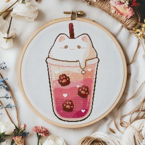 May include: A cross-stitch embroidery in a wooden hoop depicts a cat-shaped boba tea. The drink is pink and orange with brown paw prints and white hearts. The cat's head is white, and a brown straw is visible.