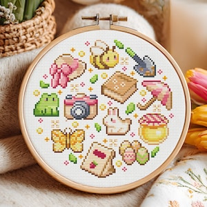 Può includere: Un telaio da ricamo a punto croce con un design a tema primaverile. Il design include un'ape, una rana, una macchina fotografica, una farfalla, un coniglio e altri elementi in uno stile colorato e pixelato. Il telaio in legno è rotondo.