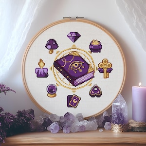 May include: A cross-stitch embroidery hoop with a central purple book design, embellished with gold detailing, encircled by magical symbols. The design includes a diamond, mushroom, pot of gold, key, and other mystical elements. The embroidery is set against a white fabric.