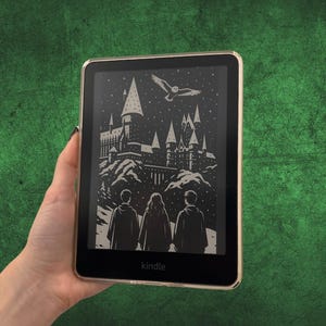 May include: A Kindle e-reader with a gold frame displays a black and white illustration of a castle, three figures, and an owl. The word "Kindle" is printed at the bottom of the screen. The e-reader is held against a green background.