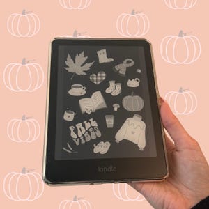 Puede incluir: Un lector electrónico Kindle negro con una pantalla que muestra varios iconos de temática otoñal, incluyendo una hoja de arce, una taza de café, un libro abierto, una calabaza, un suéter y el texto FALL VIBES en una fuente retro sobre fondo claro.
