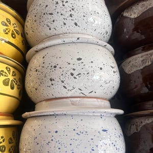 May include: A stack of ceramic pots with a speckled white glaze. The pots have a rounded shape with a wide rim and are decorated with black and blue speckles. Other pots in yellow and brown are also visible.