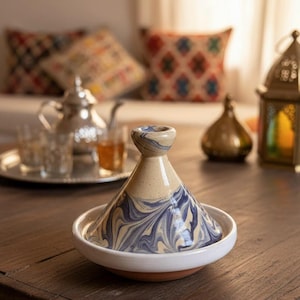 以下が含まれることがあります： クリーム色の蓋に青い渦巻き模様が施されたセラミック製のタジン鍋。ベースは白で、テラコッタの縁が付いています。鍋は濃い木製のテーブルの上に置かれ、背景にはティーセットや装飾品があります。