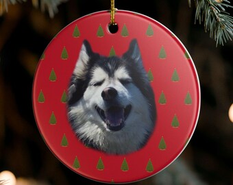 May include: A round Christmas ornament with a red background and a smiling dog's face. The ornament has a gold string for hanging and is decorated with small green Christmas trees.