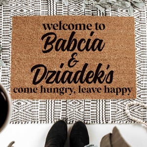 May include: A rectangular doormat made of brown coir fiber, with the words "welcome to Babcia & Dziadek's come hungry, leave happy" in black script. The mat is placed on a black and white patterned rug.