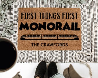 May include: A rectangular doormat made of natural coir fiber. The mat features the text "FIRST THINGS FIRST MONORAIL" in bold black letters, along with a monorail train illustration and the family name "THE CRAWFORDS".